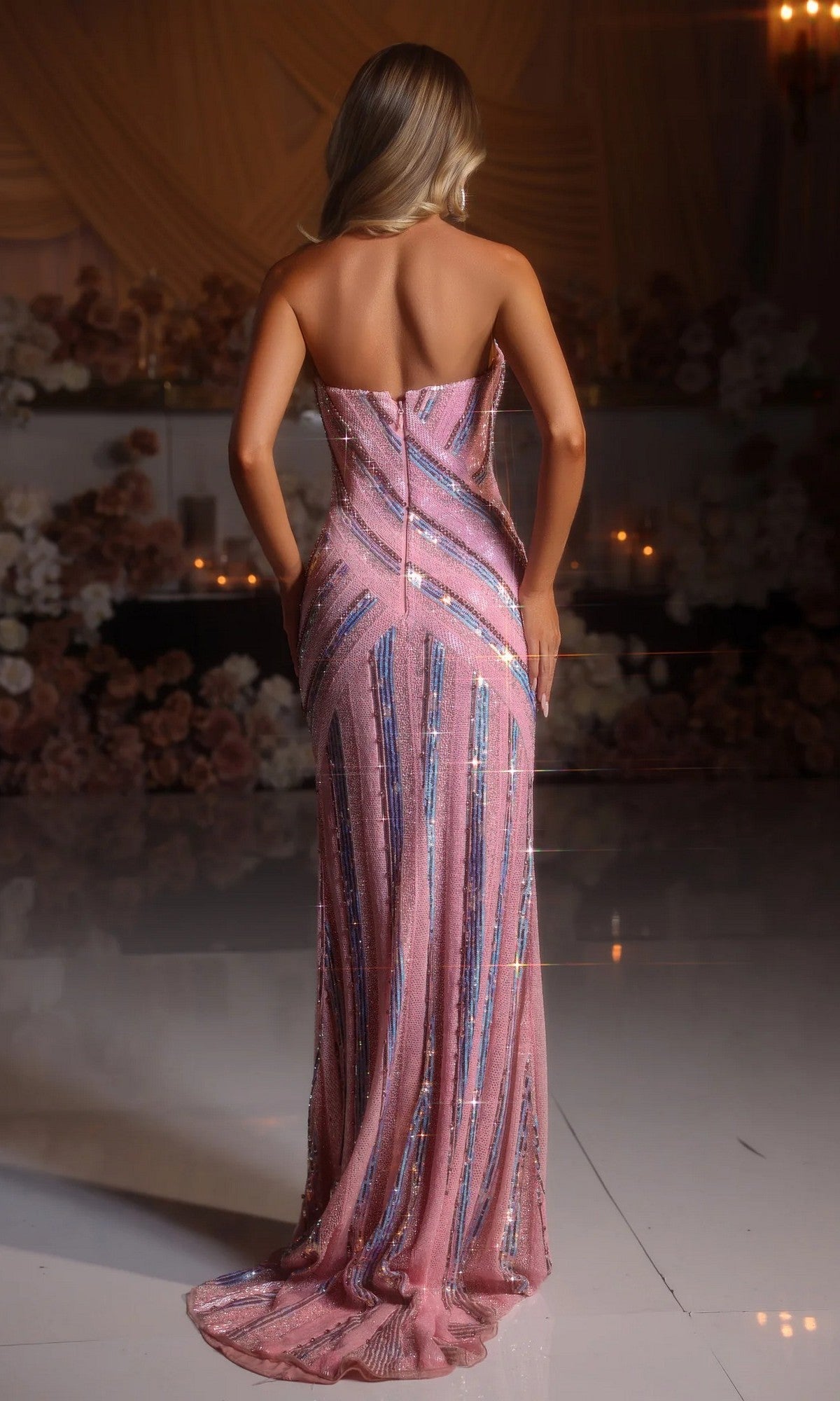 A woman with blonde hair, seen from behind, wears the Portia and Scarlett PS25449C long formal dress—a strapless pink gown with vertical sequin stripes—in an elegant, flower-adorned, dimly lit venue.